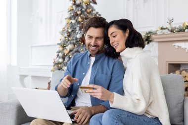 Genç bir çift, bir erkek ve bir kadın, Noel tatili için internetten alışveriş yapıyorlar. Kanepede oturup kredi kartı tutmak..