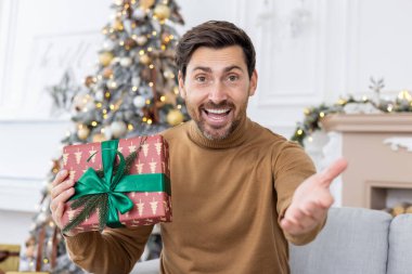 Yılbaşı ve Noel tatillerini evde kutlayan bir adam, video araması için dizüstü bilgisayar kullanıyor, webcam 'e hediye kutusunu gösteriyor, oturma odasında arkadaşlarını ve ailesini uzaktan selamlıyor..