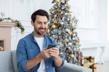 Yılbaşını ve Noel 'i evde tek başına kutlayan adam, telefondaki uygulamayı kullanarak, parti davetiyeleri göndererek, gülümseyerek, akıllı telefon tutarak, oturma odasındaki kanepede oturarak,.