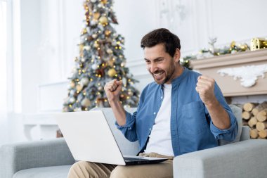 Dizüstü bilgisayara sevinçle bağıran adam internette zafer ve zafer kazandı, başarı sonuçları iyi, zaferi kutluyor, oturma odasında koltukta oturuyor, yeni yılı ve Noel 'i kutluyor..