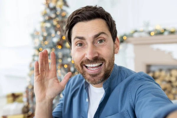 Joyful Christmas man using app on phone for online video call, man looking at smartphone camera smiling and looking at camera, waving hand greeting gesture, on sofa in living room for new year.