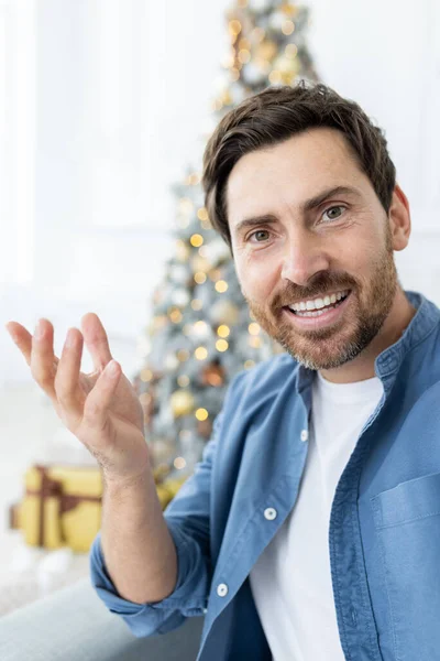 Man greets friends and family for New Year and Christmas holidays, uses app on phone, chats remotely and looks at smartphone camera, in living room on sofa near Christmas tree.