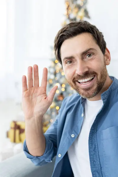 Joyful Christmas man using app on phone for online video call, man looking at smartphone camera smiling and looking at camera, waving hand greeting gesture, on sofa in living room for new year.