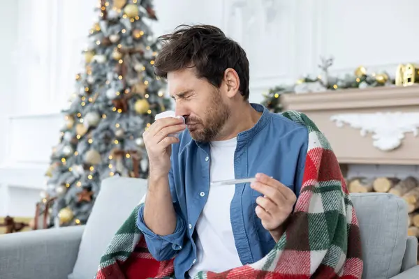 Sick and cold man sitting at home on the sofa on Christmas and New Year holidays, has a high fever covered with a blanket and sneezing in the living room of the house.