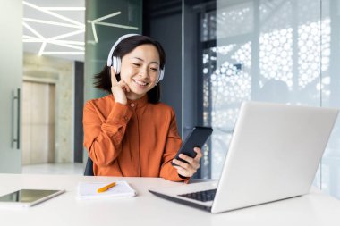 İş yerindeki genç bayan rahatlatıcı müzik dinliyor, kulaklıklı Asyalı iş kadını telefonda uygulama kullanıyor, masada dizüstü bilgisayarla oturuyor, kulaklık takıyor..
