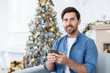 Noel ağacının yanındaki kanepede oturan ve cep telefonu kullanan genç bir adamın portresi. Kameraya bakıp gülümsüyor..