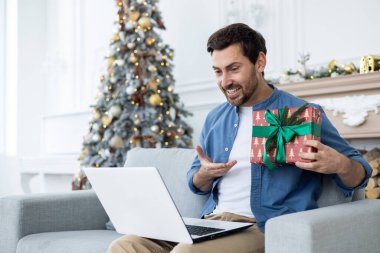 Genç bir adam internetten yeni yıl tatilini kutluyor. Evde, kanepede oturuyor ve elinde hediyesini gösteriyor..