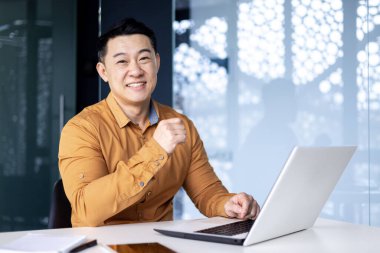İş yerindeki genç başarılı Asyalı adamın portresi, gülümseyen ve kameraya bakan adam, iş yerinde dizüstü bilgisayar kullanan, gömlekli girişimci.
