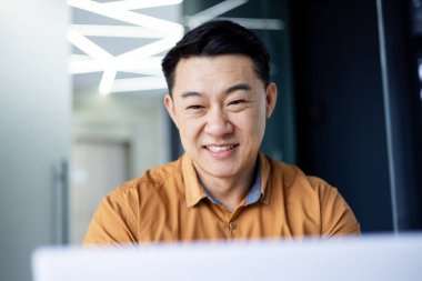 Başarılı Asyalı programcı ofis içinde dizüstü bilgisayarla çalışıyor, tişörtlü adam klavyede gülümsüyor ve daktilo kullanıyor, yeni yazılım kodluyor, işadamı başarısından memnun..