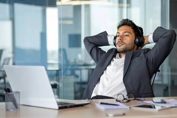 A serious thinking man looks out the window with his hands behind his head, a businessman in headphones listens to relaxing music at the workplace, thinks about solving technical financial problems.