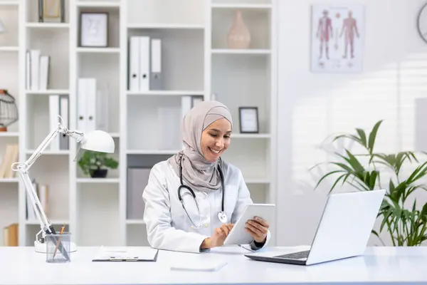 stock image Portrait Muslim female doctor in hijab, woman doctor with tablet computer using application, smiling satisfied work working inside medical office, in white medical coat, sitting at table with laptop