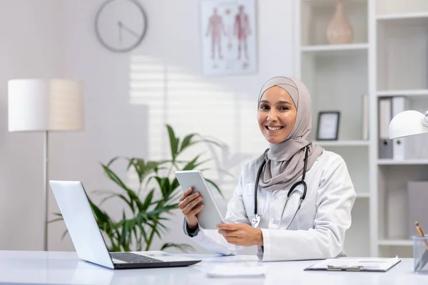 stock image Portrait of Muslim female doctor in hijab, female doctor with tablet computer looking at camera with smile, working inside medical office, wearing white medical coat, sitting at table with laptop.