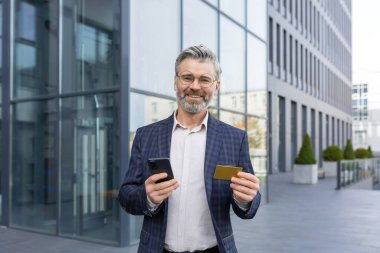 Tatmin olmuş gri saçlı bir işadamının portresi elinde telefon kartıyla online alışveriş yapıyor, modern bir ofisin önünde duruyor, satın almaktan memnun, kameraya gülümsüyor..