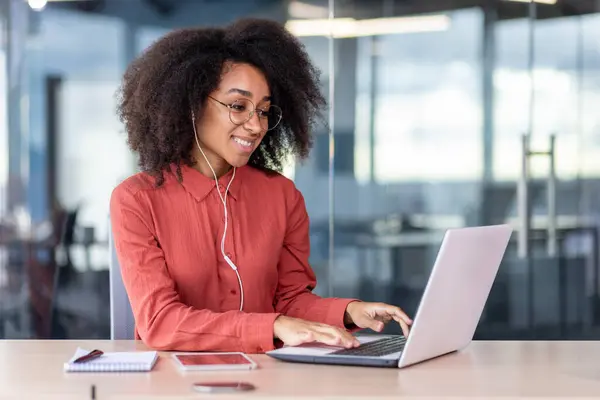 Young beautiful woman inside the office works with a laptop, a businesswoman in headphones listens music, podcasts, audio books and training course. Worker smiling , with curly hair and red shirt.