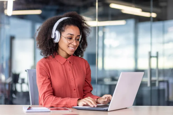 Young beautiful woman inside the office works with a laptop, a businesswoman in headphones listens music, podcasts, audio books and training course. Worker smiling , with curly hair and red shirt.