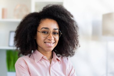 Kıvırcık saçlı güzel bir kadının portresi, Afro-Amerikan bir kadının gülümsemesi ve kameraya bakması, evdeki oturma odasında mutlu bir şekilde oturması,.