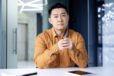 Asyalı ciddi bir ofis çalışanının portresi. Parlak bir ofiste masa başında oturmuş video görüşmesinde konuşuyor. Çinli bir adam kameraya katlanmış elleriyle bakıyor..