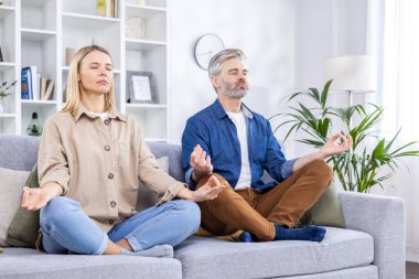 Kanepede gözleri nilüfer pozisyonunda kapalı oturan yaşlı çift oturma odasında yoga yapıyorlar. Evde spor, sağlıklı bir yaşam tarzı, yorucu bir iş gününden sonra rahatlatıcı ve rahatlatıcı..