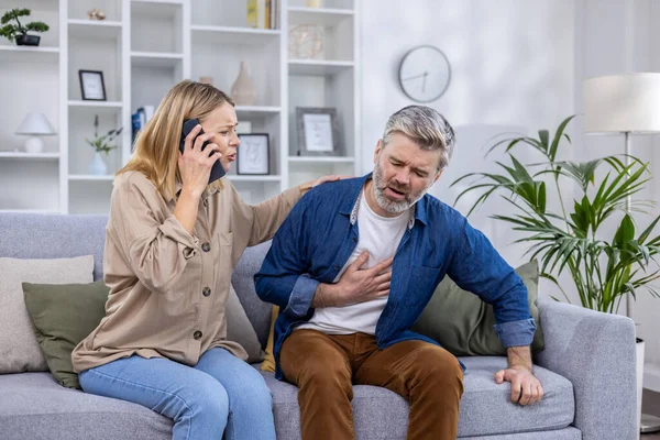 Excited scared wife is calling an ambulance on the phone because her husband is sick, he is holding his hand to his heart, bending over in pain at home, heart attack.