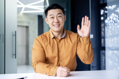 Mutlu Asyalı ofis çalışanı ışık odasında masada oturuyor ve video görüşmesinde konuşuyor Çinli adam gülümsüyor ve el sallıyor ve kameraya bakıyor..