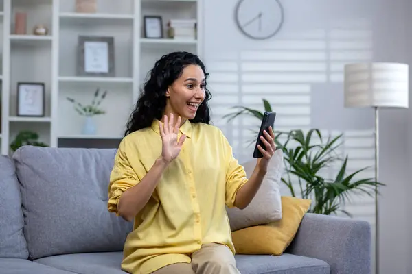 A young curly Indian woman sits on a sofa in a bright living room, holds a phone in her hand, speaks on a video call with relatives and friends, greets, smiles and waves her hand at the screen.