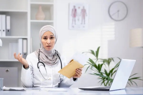 Portrait of a sad Muslim Arab female doctor sitting at a table in an office in a private clinic, holding an envelope, bad news, disappointing test results, looking at the camera.