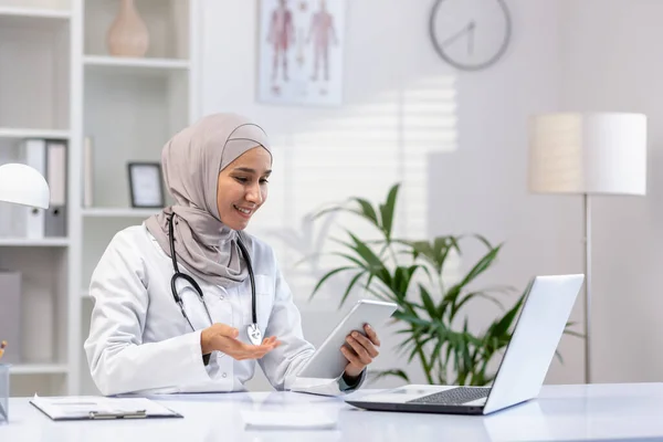 Stock image Arab Muslim woman doctor sits at a table in the office in front of a laptop, conducts an online consultation, an appointment with a patient, holds a tablet in her hands.