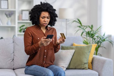 Mutsuz ve aldatılmış bir kadın oturma odasındaki kanepede oturuyor, reddedilmiş online para transferi hatası, banka kartı ve telefonu elinde tutuyor..