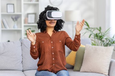 Evdeki oturma odasında yalnız başına oturan genç bir kadın sanal gerçeklik gözlüğü kullanan Afro-Amerikan bir kadın, seyahat ve uzaktan öğrenme için..