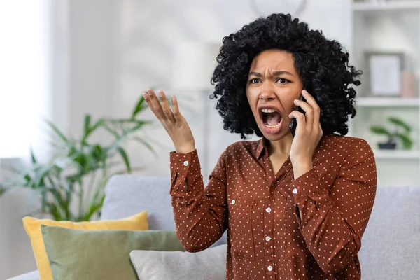 Frustrated and angry woman talking on the phone, African American woman shouting and angry at the interlocutor, sitting on the sofa in the living room of the house alone.