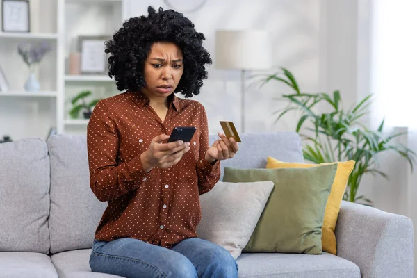 Upset unhappy and cheated woman sitting at home on sofa in living room, displeased rejected online money transfer error, holding bank credit card and phone.