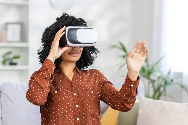 Young woman alone sitting on couch in living room at home, African American woman using virtual reality glasses, for travel, and remote learning.