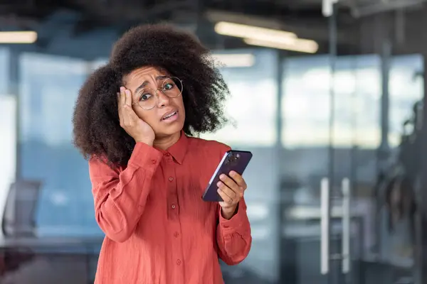 Portrait of dissatisfied sad disappointed woman inside office, businesswoman with phone in hands looking at camera confused and nervous, standing near window.