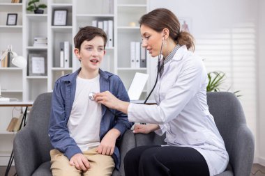 Bir kadın doktorla randevusu olan genç bir çocuk özel bir klinikteki bir ofiste sandalyeye oturur. Bir çocuk doktoru steteskobu olan bir çocuğu muayene eder..