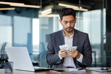 Ciddi Arap işadamı, modern bir ofiste dizüstü bilgisayarın önündeki masada oturuyor. Kazandığı parayı, online kazancını, elinde bir tomar doları sayıyor..