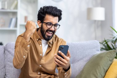 Evdeki kazananın portresi, mutlu mutlu adam çevrimiçi bildiri kazanma, telefonu tutma, zafer jesti, başarı sonuçlarından tatmin olma, oturma odasında kanepede oturma..