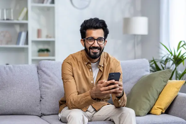 Portrait of a young man at home, Indian man sitting on the sofa in the living room of the house, smiling, looking at the camera, holding a phone, using an application on a smartphone, typing a message