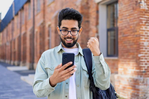 Young indian student received online win notification message phone, successful entrance exam results, man with backpack walking outside university campus, celebrating successful achievement result.