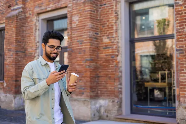 Young man walking in the city reading messages on the phone smiling, with a cup of hot drink, businessman entrepreneur programmer in casual clothes using an application on a smartphone.