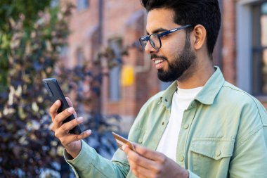 Ofis binasının dışındaki genç adam bankta oturuyor ve şehirde yürüyor, telefonu internetten satın almak için kullanıyor, iş adamı banka kredi kartını tutuyor gülümsüyor, günlük giysiler giyiyor..