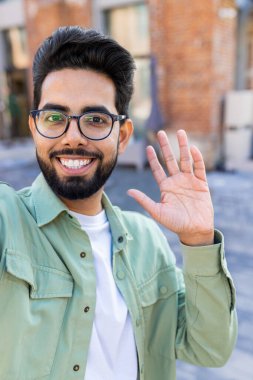 Telefon web kamerası görüntüsü, akıllı telefondaki uygulamayı uzaktan iletişim için kullanıyor, Hintli adam gülümsüyor ve kameraya bakıyor, el sallıyor. Dikey çekim.
