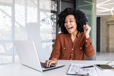 Joyful successful satisfied african american businesswoman celebrating great achievement results at workplace inside office, female worker celebrating holding hand up smiling with laptop. clipart