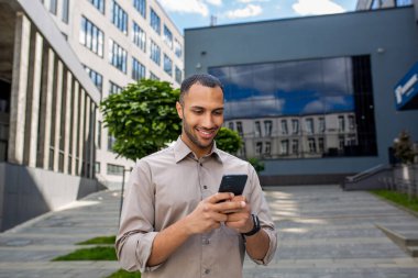 Tişörtlü başarılı bir erkek işadamı ofis binasının dışında yürüyor, elinde bir telefon tutuyor, akıllı telefondan başvuru yapıyor, online sayfalar okuyor, mutlu bir şekilde gülümsüyor.