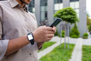 Erkek eli kapalı, gömlekli iş adamı elinde telefon, akıllı telefon uygulaması kullanıyor, ofis binasının dışında yürüyor..