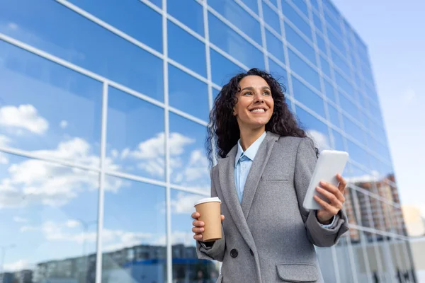 Successful Hispanic businesswoman walks to the office, holds a coffee and a phone in her hands, smiles and solves cases on the way to work, stands on the street of the office.