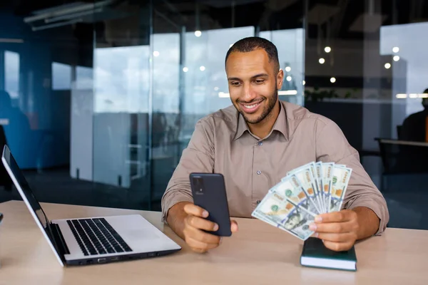 Successful businessman sitting at the table in the office in front of the laptop, holding the phone and dollars in his hands, rejoices at the win, looks into the window of the smartphone.