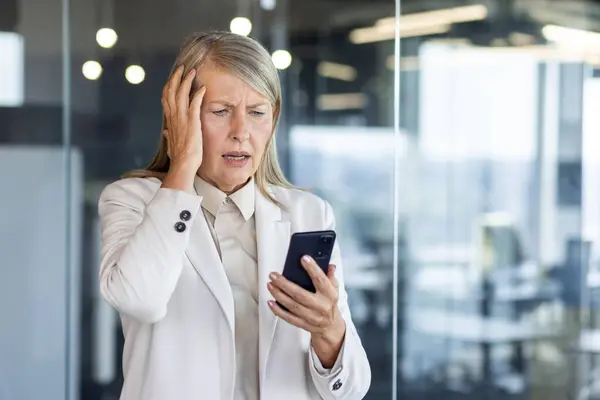 Senior mature woman boss at workplace received online notification message on phone with bad news, financier disappointed sad standing near window inside office, using smartphone.