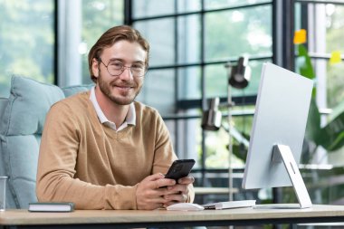 Gözlüklü arkadaş canlısı iş adamı masadaki ofiste çalışıyor, telefonu elinde tutuyor, mesaj atıyor, gülümsüyor ve kameraya bakıyor..