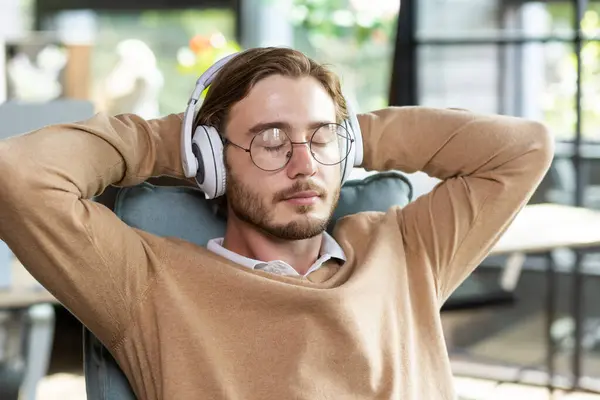 Portrait of a tired office worker in headphones resting at the workplace in a chair, arms crossed on the title with closed eyes, napping during a break between work, listening to music.