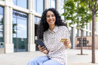 Kıvırcık saçlı güzel bir kadının portresi, ofis binasının dışında bir bankta oturuyor, gülümsüyor ve kameraya bakıyor, elinde telefon, banka kartı, online servis siparişi veriyor, alışveriş yapıyor..